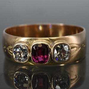 May include: A gold ring with three gemstones. The center stone is a deep red, flanked by two clear, square-cut stones. The ring's band is wide and features subtle engraved details. The ring is displayed on a reflective surface.