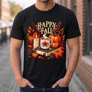 May include: A black t-shirt featuring a graphic print with the text HAPPY FALL. The design includes a steaming mug, a lit candle, pumpkins, pinecones, and autumn leaves arranged on top of a closed book, all rendered in warm orange and red tones.
