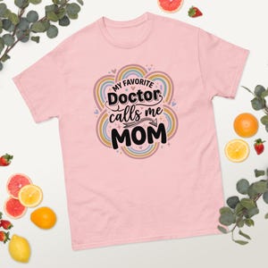 以下が含まれることがあります： 「My Favorite Doctor calls me MOM」の文字がカラフルなデザインでプリントされた、薄いピンク色のTシャツ。デザインには虹色のアーチとハートが含まれています。このシャツは半袖のカジュアルスタイルです。