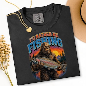 Può includere: T-shirt nero con la scritta "I'd Rather Be Fishing" e una grafica di Bigfoot che tiene una trota iridea. Il design include un tramonto, alberi e acqua. La maglietta ha un'etichetta Comfort Colors, taglia XL.