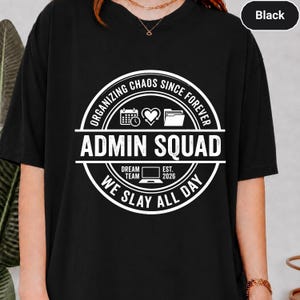 May include: A black t-shirt featuring a white circular graphic with the text "ORGANIZING CHAOS SINCE FOREVER", "ADMIN SQUAD", "DREAM TEAM", "EST. 2026", and "WE SLAY ALL DAY". The design includes icons of a calendar, a heart, a file folder, and a laptop.