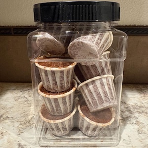 May include: A clear plastic square jar with a black screw-top lid containing multiple small, round, brown baked goods in white fluted paper liners. The container is filled with several stacked individual portions of the dark brown food items.