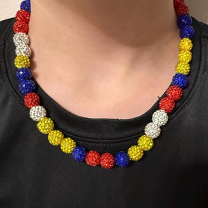 May include: A colorful beaded necklace featuring red, white, yellow, and blue spherical beads. The necklace is worn against a black background, highlighting the vibrant colors of the beads. The beads are evenly spaced, creating a visually appealing accessory.