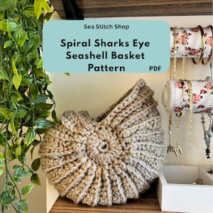 Pode incluir: Uma cesta de concha marinha de crochê em espiral, feita com fio bege. A imagem inclui o texto "Sea Stitch Shop" e "Spiral Sharks Eye Seashell Basket Pattern PDF". A cesta está em uma prateleira de madeira.