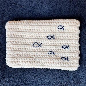 May include: Cream-colored, rectangular crochet item with a textured surface. Features several small, embroidered blue fish scattered across the surface. The item is likely a decorative piece or a small mat.