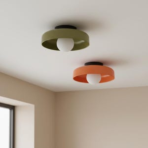 May include: Two modern ceiling lights. One is olive green, the other is orange. Each has a black base and a white globe bulb. The lights are mounted on a white ceiling in a room with beige walls.