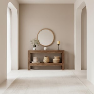 Puede incluir: Una consola de madera con un espejo redondo, objetos decorativos y una alfombra. La mesa tiene dos estantes y está adornada con un jarrón, una vela y cestas tejidas. La habitación tiene una paleta de colores neutros con arcos.