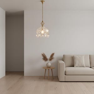 May include: A clear glass pendant light with a brass finish hangs from the ceiling. The light fixture has a decorative, bell-shaped shade with crystal accents. The room features a neutral colour palette with a beige sofa and light wood flooring.