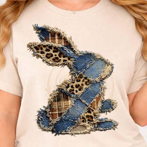 Puede incluir: Una camiseta beige con un diseño de conejo de patchwork. El conejo está compuesto de mezclilla, estampado de leopardo y telas a cuadros. El diseño tiene un borde deshilachado y desgastado, lo que le da un aspecto rústico. Es una prenda casual para el día a día.