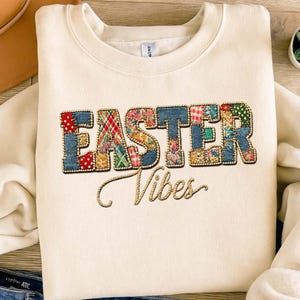 May include: Cream-colored sweatshirt with the words "EASTER Vibes" embroidered on the front. The word "EASTER" is in a patchwork design with denim and floral patterns. The word "Vibes" is in a cursive font.