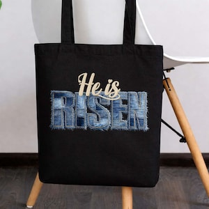 Puede incluir: Bolso tote negro con la frase "He is Risen" en un diseño estilo denim. El texto está en una fuente de escritura y está centrado en la bolsa. La bolsa tiene asas negras y está colgada de una silla.