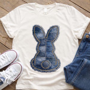 Puede incluir: Camiseta blanca con un diseño de conejo estilo denim. El conejo está hecho de parches de mezclilla azul con un contorno cosido amarillo. Un par de zapatillas blancas y unos vaqueros azules también están en la imagen.