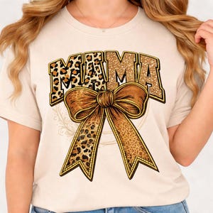 Puede incluir: Camiseta beige con la palabra "MAMA" en purpurina dorada y estampado de leopardo. Un gran lazo marrón con un diseño de estampado de leopardo está centrado debajo del texto. La camiseta es de estilo casual.