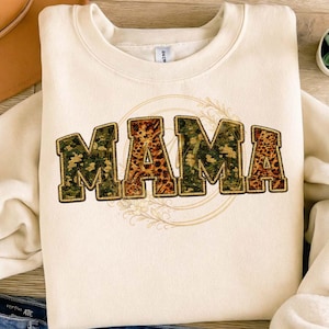 Puede incluir: Sudadera de color crema con la palabra "MAMA" en un diseño de letras en negrita. Las letras presentan un estampado de camuflaje y leopardo. Un diseño floral rodea la palabra. La sudadera está doblada sobre una superficie de madera.