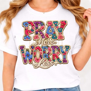 Puede incluir: Camiseta blanca con el texto "PRAY More WORRY Less" en un diseño floral. El texto tiene un contorno dorado y está relleno de flores rosas, azules y moradas.