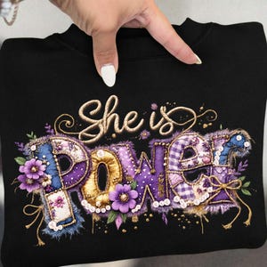 Puede incluir: Sudadera negra con las palabras "She is POWER" en un diseño decorativo. La palabra "POWER" está en morado, dorado y azul con detalles florales y de perlas. Una mano sostiene la sudadera.