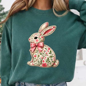 Puede incluir: Una sudadera verde azulado con un diseño de conejito floral. El conejito tiene un lazo rosa y un patrón de flores rosas y hojas verdes. El diseño está centrado en la parte delantera de la sudadera.