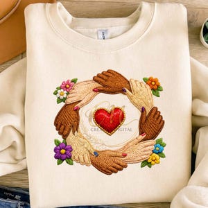 Puede incluir: Sudadera color crema con un diseño bordado. El diseño presenta manos en círculo alrededor de un corazón rojo, con flores coloridas. También está la palabra "DIGITAL". La sudadera está hecha de un material suave.