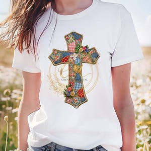 Może przedstawiać: Biała koszulka z kolorowym wzorem krzyża. Krzyż zdobiony jest kwiatowymi i patchworkowymi wzorami, złotymi akcentami oraz napisem "CREATIONS TAL". Projekt jest umieszczony centralnie na koszulce.