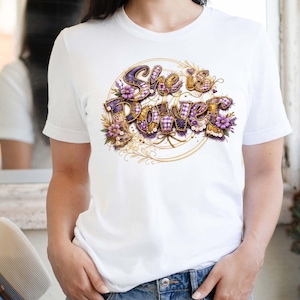 Puede incluir: Camiseta blanca con el texto "She is Power" en un diseño decorativo. El texto es en morado y dorado con detalles florales. La camiseta es de cuello redondo y tiene mangas cortas.