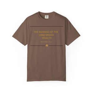 Puede incluir: Camiseta marrón con el texto dorado "THE BLESSING OF THE LORD BRINGS WEALTH. PROVERBS 10:22". El diseño incluye líneas horizontales y un icono de llama. Una prenda casual para el uso diario.