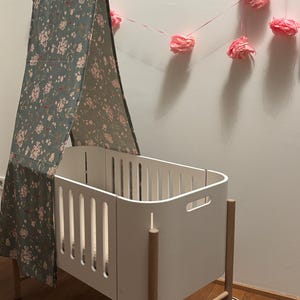 Può includere: Una culla bianca con una tenda da sole con un motivo floreale rosa e verde. La culla ha gambe in legno e una ruota su un lato. Fiori di carta rosa sono appesi sullo sfondo.