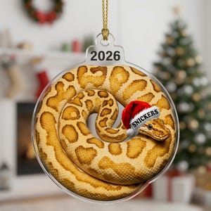May include: A clear, circular acrylic Christmas ornament featuring a coiled yellow and brown patterned snake wearing a red and white Santa hat with the name SNICKERS printed on the brim. The year 2026 is printed in black at the top of the ornament.