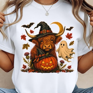 May include: A white t-shirt featuring a graphic print of a brown Highland cow wearing a black witch hat, positioned above a jack-o'-lantern with a small witch hat. A beige ghost, bats, a crescent moon, and autumn leaves surround the central figures.