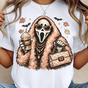Peut inclure: Un t-shirt blanc avec une illustration d'un personnage fantôme portant un manteau en fausse fourrure beige, des lunettes de soleil œil-de-chat et des colliers. Il tient un café à la citrouille et un sac matelassé, entouré de feuilles et de chauves-souris.