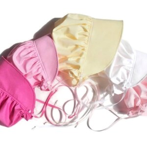 This is a photo of 5 prairie style bonnets with the wide brim facing right. They are overlapping on an angle. From left to right they are bright pink, medium pink, yellow, white, light pink.  The ribbon ties are in the middle of the photo.