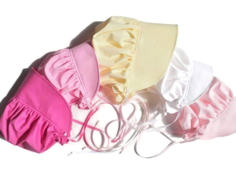 This is a photo of 5 prairie style bonnets with the wide brim facing right. They are overlapping on an angle. From left to right they are bright pink, medium pink, yellow, white, light pink.  The ribbon ties are in the middle of the photo.