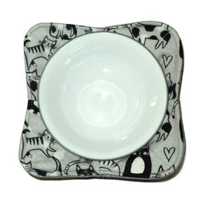 May include: A grey and white cat-themed bowl mat with a white ceramic bowl on top. The mat features a repeating pattern of black and white cats and hearts.