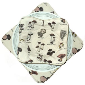 May include: Two beige fabric pot holders with a mushroom print and the names of the mushrooms. The pot holders are on top of a white plate with a blue rim.