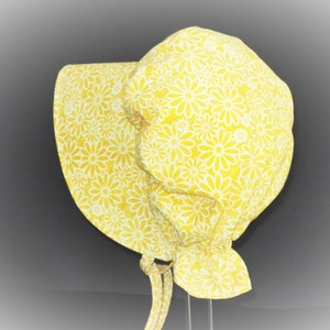 Wide brim prairie bonnet facing left. Pretty bright yellow floral fabric with white etched daisy flowers packed together with no leaves or stems. Fabric is ruffled at bottom back, and fabric ties are looped at bottom of brim.