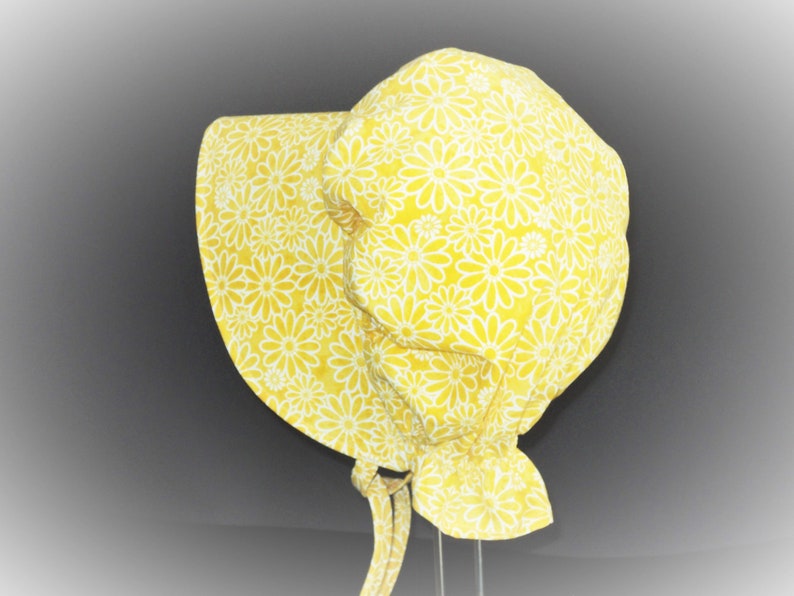 Wide brim prairie bonnet facing left. Pretty bright yellow floral fabric with white etched daisy flowers packed together with no leaves or stems. Fabric is ruffled at bottom back, and fabric ties are looped at bottom of brim.