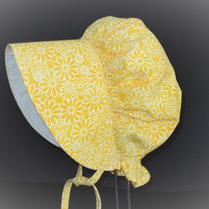 This is a pioneer/prairie style bonnet with a large brim in a floral print. There are  sketched white daisies on a bright yellow background. There is elastic at the back of the neck. The brim is white on the inside. It has matching fabric ties.