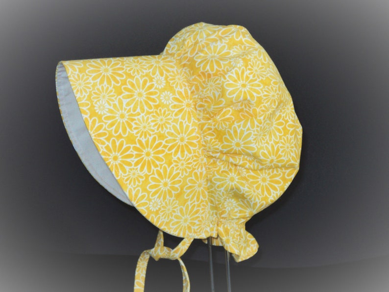 This is a pioneer/prairie style bonnet with a large brim in a floral print. There are  sketched white daisies on a bright yellow background. There is elastic at the back of the neck. The brim is white on the inside. It has matching fabric ties.