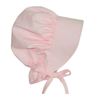 Prairie bonnet with wide brim facing right. It is solid light pink. The side-back is puffy and gathered onto the brim. There is a gathered ruffle at the bottom back. A pink bow is tied under the hat with ties hanging down.