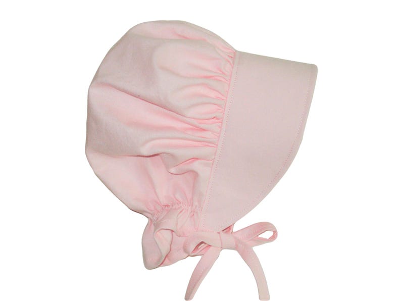 Prairie bonnet with wide brim facing right. It is solid light pink. The side-back is puffy and gathered onto the brim. There is a gathered ruffle at the bottom back. A pink bow is tied under the hat with ties hanging down.