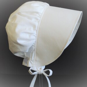 Prairie bonnet with wide brim facing right. It is solid white. The side-back is puffy and gathered onto the brim. There is a gathered ruffle at the bottom back. A white bow is tied under the hat with ties hanging down.