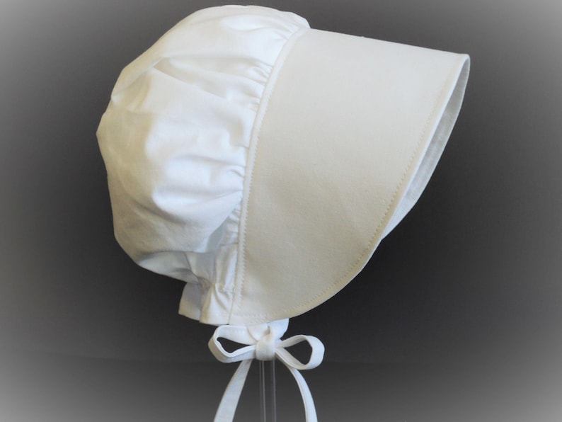 Prairie bonnet with wide brim facing right. It is solid white. The side-back is puffy and gathered onto the brim. There is a gathered ruffle at the bottom back. A white bow is tied under the hat with ties hanging down.