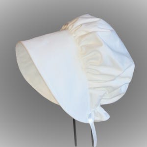 Prairie bonnet with wide brim facing left. It is solid white. The side-back is puffy and gathered onto the brim. There is a gathered ruffle at the bottom back. A white ribbon tie hangs straight down.