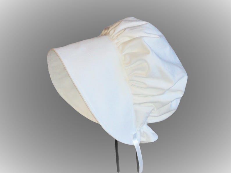 Prairie bonnet with wide brim facing left. It is solid white. The side-back is puffy and gathered onto the brim. There is a gathered ruffle at the bottom back. A white ribbon tie hangs straight down.