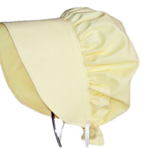 Prairie bonnet with wide brim facing left. It is solid light yellow. The side-back is puffy and gathered onto the brim. There is a gathered ruffle at the bottom Natural ribbon ties hang down.