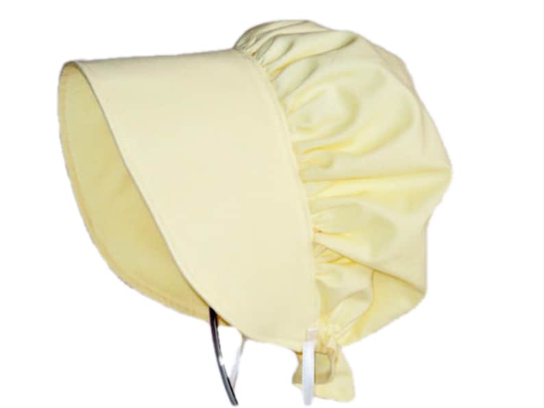 Prairie bonnet with wide brim facing left. It is solid light yellow. The side-back is puffy and gathered onto the brim. There is a gathered ruffle at the bottom Natural ribbon ties hang down.