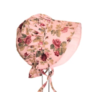 May include: A pink sun hat with a floral pattern of roses. The hat has a ruffle around the brim and ties at the back.