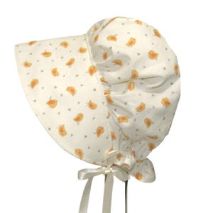 May include: A cream-coloured baby bonnet with a brim and a gathered crown. The fabric is patterned with small orange birds and tiny grey dots. A white satin ribbon is tied under the chin. The bonnet is designed for infants and toddlers.