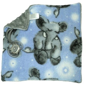 May include: A blue baby blanket with a grey fleece backing. The blanket features a repeating pattern of grey elephants on a light blue background with white dots. A grey pacifier is attached to the blanket with a grey ribbon.