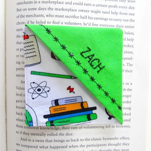 May include: A green fabric bookmark with the name "ZACH" in black letters. The bookmark is triangular and has a patterned fabric on the bottom with school-themed illustrations. It is placed inside an open book.