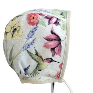 Peut inclure: Un bonnet pour bébé à motifs floraux avec un colibri, de la lavande et diverses fleurs dans les tons de rose, jaune et violet. Le bonnet a une bordure beige et des liens.
